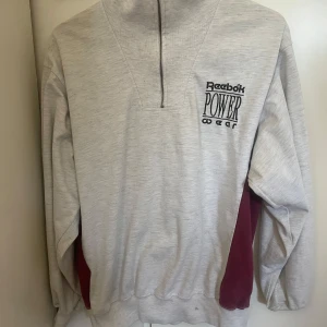 Grå Reebok Power Wear half zip tröja - Säljer en grå långärmad tröja från Reebok Power Wear med half zip och coolt broderat tryck på bröstet. Tröjan har vinröda paneler på sidorna och är tillverkad i mjuk bomullsmix. Perfekt för träning eller chill dagar. Oversized passform för skön vibe. Den är använd ganska mycket och har därför lite fläckar ( kolla på bild) 