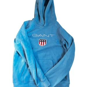 Blå hoodie från GANT med logga - Blå hoodie från GANT med broderad logga och sköld på bröstet. Tröjan har huva, magficka och långa ärmar. Tillverkad i mjuk bomull, perfekt för chill dagar. Snyggt sportig look med klassisk GANT-stil.