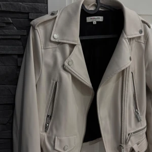 Vit skinnjacka från Morgan, strl 36 - Snygg vit skinnjacka från Morgan med coola silvriga dragkedjor och detaljer. Jackan har klassisk bikerstil med bred krage, bälte i midjan och flera fickor med dragkedja. 