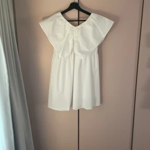 Vit kortklänning med volang från Zara - Supersöt vit kortklänning från Zara med stor volangkrage och rynkade detaljer framtill. Klänningen har en loose passform och är tillverkad i mjuk bomull. Perfekt för dig som gillar en clean och trendig look med lite extra statement vid axlarna.