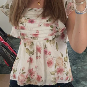 Blommig vit blus med volanger - Blus från MQ. Supersöt blommig blus i vit med rosa och beige blommor, gröna blad och små prickar. Blusen har breda volangärmar, fyrkantig halsringning och smock i ryggen för skön passform. Materialet är lätt och luftigt, perfekt för varma dagar.