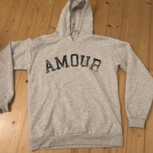 Grå hoodie med tryck från Gina Tricot - Snygg ljusgrå hoodie från Gina Tricot med svart AMOUR-tryck på bröstet och en cool stjärna på ryggen. Tröjan har huva med snörning och långa ärmar. Perfekt för chill dagar och streetstyle. Mjuk bomullsmix som känns skön mot huden.