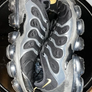 Nike Air Vapormax Plus svart/guld - Nike Air Vapormax Plus sneakers i svart med detaljer i guld och grått. Snyggt vågigt mönster på ovansidan och genomskinlig, bubblig sula som ger en futuristisk vibe. Skorna har snörning och är tillverkade i syntet och textil. Perfekt för dig som gillar streetstyle och vill sticka ut.