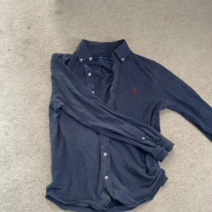 Blå långärmad skjorta från Ralph Lauren - Snygg mörkblå skjorta från Ralph Lauren med klassisk krage och knappar framtill. Lätt mesh-material som andas och har en röd broderad logga på bröstet. Perfekt för dig som gillar stilrena och tidlösa plagg med en sportig touch.