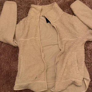 vit teddyjacka från 157 XS - Mysig vit teddyjacka från 157 i storlek XS. Jackan har lång ärm, hög krage och dragkedja framtill. Perfekt för kyliga dagar och enkel att slänga på sig när man vill vara extra comfy. Fleece-materialet gör den supermjuk och varm.