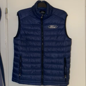 Blå dunväst från Southwest x Ford - Snygg marinblå dunväst från Southwest med Ford-logga broderad på bröstet. Västen har ståkrage, två sidofickor med dragkedja och quiltad design. Perfekt för dig som gillar bilar och vill ha en cool look med sportig vibe.