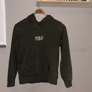 Mörkgrön hoodie från Polo Ralph Lauren - Snygg mörkgrön hoodie från Polo Ralph Lauren med vit logga på bröstet. Tröjan har huva, lång ärm och en klassisk magficka framtill. Perfekt för chill dagar och enkel att matcha med jeans eller joggers. Materialet är mjuk bomull som känns skönt mot huden. Tröjan är i väldigt bra skick och har inte använts många gånger. Perfekt grön färg inför hösten🍂🍁.🍀PRIS KAN DIGITALISERAS VID EN SNABB AFFÄR 🍀