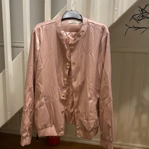 Rosa satin bomberjacka från Daphnea - Snygg rosa bomberjacka i satin från Daphnea Paris med guldfärgade knappar och broderade fåglar på ryggen. Jackan har rund hals, ribbade muddar och fickor framtill. Perfekt för dig som vill sticka ut med en unik och glansig look. Frakt ej inklusive.