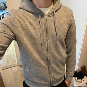 Grå hoodie från Band of Brothers - Säljer en grå hoodie från Band of Brothers med dragkedja och snörning i huvan. Tröjan har ribbade muddar, två fickor fram och är tillverkad i mjuk bomullsmix. Perfekt för chill dagar eller när du vill ha något bekvämt och stilrent.