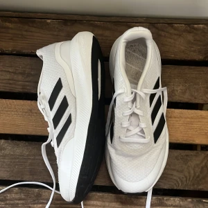 Vita Adidas sneakers med svarta ränder - Snygga vita Adidas sneakers med klassiska svarta ränder på sidorna. Skorna har en rund tå, platt sula och snörning framtill. Ovandelen är i mesh och syntetmaterial för bra andningsförmåga. Perfekta för dig som gillar sportig stil och vill ha bekväma skor till vardagen.
