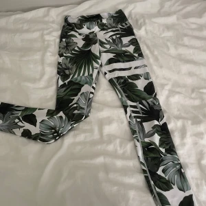 Tropiska leggings med bladmönster - Snygga leggings med tropiskt bladmönster i grönt, svart och grått på vit botten. De har en tight passform och är perfekta för träning eller chill. Materialet är mjukt och stretchigt, vilket gör dem riktigt bekväma att bära. Midjan är normal och de sitter skönt hela dagen.
