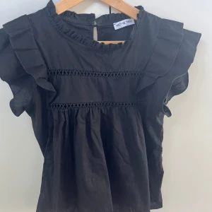 Svart blus med volang från Meet Me There - Svart blus med volangdetaljer på axlarna och rynkad nederdel. Blusen har rund hals med liten volangkant. Den är i väldigt bra skick då jag inte använt den så mycket.