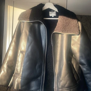 Svart teddyjacka från Global Funk - Säljer en svart teddyjacka från Global Funk med mjukt fuskpälsfoder och bred krage. Jackan har en snygg skinnlook på utsidan, dragkedja framtill och två fickor. Perfekt för dig som vill ha en cool och varm jacka med lite oversized känsla.