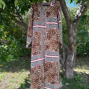 Långklänning med leopardmönster och knappar - Snygg långklänning med leopardmönster och grafiska detaljer i svart, vitt och rött. Klänningen har krage, långa ärmar och knappar framtill. Materialet känns lätt och mjukt, perfekt för dig som gillar att sticka ut med djurmönster och coola kontraster. Storlek XL. Kanske passar runt en L/XL. Myclet fint skick, men tyvärr finns inte midjebandet kvar. Samt se bilder på knapphålet att det kan vara någon lös tråd. 