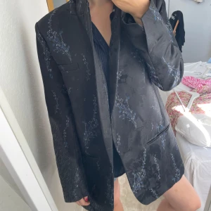 Svart blommig kavaj H&M Conscious - Snygg svart kavaj från H&M Conscious Exclusive med diskret blommigt mönster i blått. Oversized passform med klassisk krage och fickor fram. Materialet har en lätt glansig finish som ger en lyxig känsla.