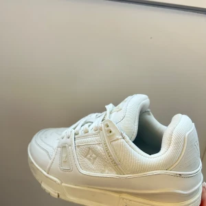 Louis Vuitton vita sneakers i skinn - Säljer ett par fräscha vita Louis Vuitton sneakers med klassisk LV-logga på sidan och tungan. Skorna har snörning, rund tå och platt sula. Tillverkade i lyxigt skinn med detaljer i textil och präglade mönster. Perfekt för dig som gillar exklusiv streetstyle. Tagit på mig de en gång bara, kommer med box och dustbag 