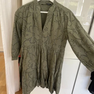 Grön kortklänning med broderade detaljer - Säljer en grön kortklänning från Zara med broderade blommönster och hålbroderier. Klänningen har v-ringning, knappar framtill och långa ärmar. Tillverkad i mjuk bomull med volang nedtill för en chill och feminin vibe. Klänning är i mycket bra skick och använd endast en gång. 