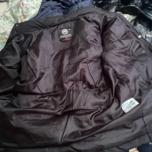 Svart pufferjväst  från Canada Goose - Säljer en svart pufferjacka från Canada Goose med klassisk logga på bröstet och stora fickor framtill. Jackan har en hög krage och knäppning med både dragkedja och tryckknappar. Materialet är polyester och dun, perfekt för kalla dagar. köpte den från plick passar inte mig den är lite trasig 