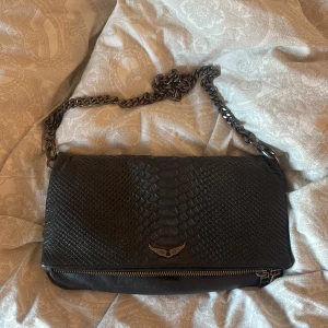 Svart axelväska från Zadig & Voltaire - Snygg svart axelväska äkta från Zadig & Voltaire i skinn med reptilpräglad yta och grov kedja som axelrem. Väskan har metalldetaljer, dragkedja både fram och bak samt en liten emblem med vingar på framsidan. Perfekt för dig som gillar edgy och stilren design. Köpte den på Plick men har aldrig kommit till användning. Kedjan o andra små detaljer har tappat färg därför säljer jag den billigt💕Påse medföljer