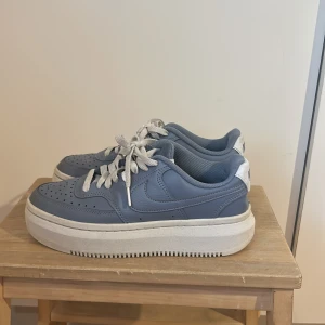 Nike Air Force 1 Sage blå - Nike Air Force 1 sneakers i en snygg blå färg med vita detaljer och snörning. Skorna har en platt, tjock sula och perforerad tå för extra ventilation. Tillverkade i skinn med meshfoder för skön komfort. Perfekt för dig som gillar klassisk streetstil och vill sticka ut.