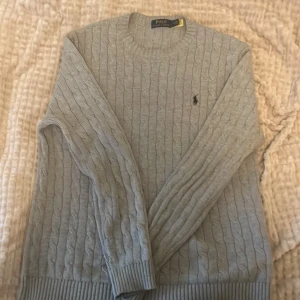 Grå kabelstickad tröja Polo Ralph Lauren - Snygg grå kabelstickad tröja från Polo Ralph Lauren i storlek L. Tröjan har rund hals, långa ärmar och den klassiska broderade loggan på bröstet. Perfekt för dig som gillar stilrena och tidlösa plagg med premiumkänsla. Vid snabbt köp kan pris diskuteras! 