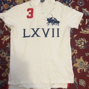 Vit Polo Ralph Lauren Polo - Vit polo från Polo Ralph Lauren med klassisk krage och korta ärmar. Stor röd siffra 3 på både fram- och baksidan, samt mörkblå polospelare och LXVII-tryck på bröstet. Custom fit i mjuk bomull. Inga fläckar som ny.