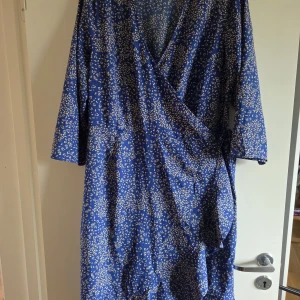 Blå omlottklänning från Vero Moda - Snygg blå långklänning från Vero Moda med ljusblommigt mönster. Klänningen har omlottmodell med v-ringning, trekvartsärm och knyt i midjan. Materialet är mjukt och lätt, perfekt för vår och sommar. Passformen är normal och den har en feminin siluett.