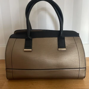 Svart och guld handväska med dragkedja - Snygg handväska i svart och guld med stilrena detaljer och metallfästen. Väskan har två rejäla handtag och en praktisk dragkedja upptill. Materialet är skinnimitation och insidan är fodrad. Perfekt storlek för att få plats med allt du behöver.