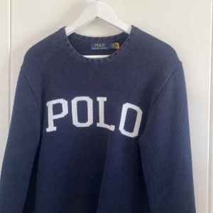 Mörkblå stickad tröja från Polo Ralph Lauren - Snygg mörkblå stickad tröja från Polo Ralph Lauren med stor vit POLO-text framtill och klassisk logga broderad vid nederkanten. Tröjan har rund hals och långa ärmar, perfekt för dig som gillar stilren och sportig vibe.