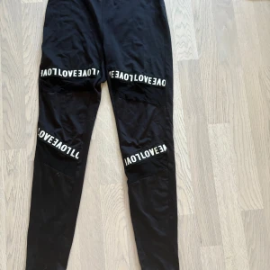 Svarta leggings/träningsbyxor - Svarta leggings i storlek S med vita textdetaljer 'LOVE' på båda benen. Materialet är mjukt och stretchigt, med meshpaneler vid texten för extra cool look. Perfekta för chill eller träning, med slim passform och elastisk midja.