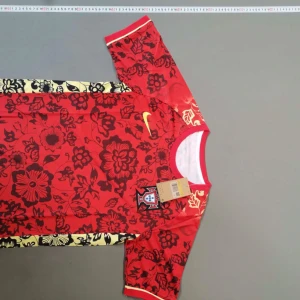 Portugal röd blommig Nike fotbollströja - Snygg Portugal fotbollströja från Nike i rött med svarta och gula blommor över hela tröjan. Kortärmad med rund hals och Portugal-emblem på bröstet. Materialet är lätt och andas, perfekt för match eller träning. Gult Nike-swoosh på framsidan.