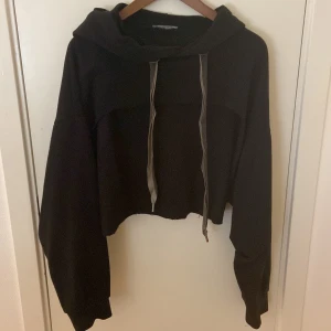Svart croppad hoodie med snörning - Svart croppad hoodie med långa ärmar och bred huva. Tröjan har coola snören framtill och en loose passform som ger en avslappnad vibe. Perfekt för dig som gillar streetstyle och vill ha något bekvämt men trendigt.