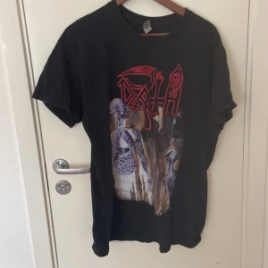 Death Human band t-shirt svart L - Svart t-shirt från Gildan med tryck från Deaths album Human. Framsidan har ett stort, färgstarkt motiv med röda och lila detaljer och bandets logga, baksidan har HUMAN i rött och albumspår i cirkel. Klassisk passform och rund hals. Perfekt för metalheads.