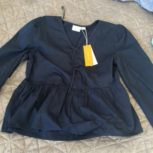 Mörkblå blus med knyt från Gina tricot  - Säljer en mörkblå blus från gina tricot t med v-ringning och knyt framtill. Blusen har långa ärmar och en volang nedtill som ger en cool och feminin vibe. Tillverkad i mjuk bomull, perfekt för dig som gillar stilrena och bekväma plagg.