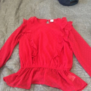 Röd blus med volanger och prickar - Säljer en röd blus från Lindex  med volanger längs axlarna och framtill. Blusen har ett diskret prickigt mönster, långärmade transparenta ärmar och en lätt peplum-effekt vid midjan. Perfekt för dig som gillar färg och detaljer!