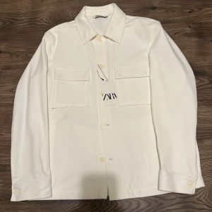 Vit overshirt från Zara XL - Snygg vit overshirt från Zara i storlek XL. Jackan har klassisk krage, två stora bröstfickor med lock och knäppning framtill med matchande vita knappar. Materialet är mjukt och känns som bomull, perfekt för lager-på-lager. Stilren och clean look.   OBS! Jackan har aldrig använts 