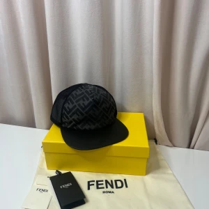 Fendi keps - Snygg svart keps från Fendi med ikoniskt FF-mönster framtill och mesh på sidorna och bak. Justerbar rem med gulddetalj och Fendi-logga. Skärmen är böjd och kepsen är tillverkad i polyester och bomull för en lätt och luftig känsla.