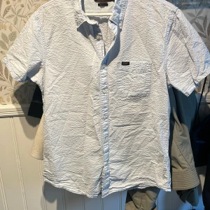 Vit kortärmad skjorta från Lee - Fräsch vit skjorta från Lee med korta ärmar och regular fit. Skjortan har en strukturerad yta, bröstficka med Lee-logga och knäppning framtill. Perfekt för varma dagar och enkel att styla till jeans eller shorts.