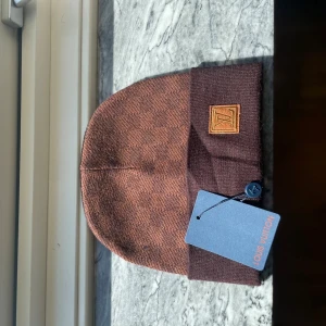 Brun rutig mössa från Louis Vuitton - Snygg brun mössa från Louis Vuitton med klassiskt rutigt mönster och LV-logga på uppvikt kant. Mössan är stickad och har en exklusiv känsla, perfekt för dig som vill ha något extra. Taggen sitter kvar och märket syns tydligt på insidan.