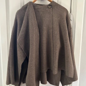 Brun oversized kofta från Zara - Säljer en brun oversized jacka/cape från Zara med cool asymmetrisk design och knappdetalj vid halsen. Den har långa ärmar och är tillverkad i mjukt stickat material som ger en chill och avslappnad vibe. Perfekt för lager-på-lager och att mysa i under kyliga dagar. Använd mindre än 5 gånger. 