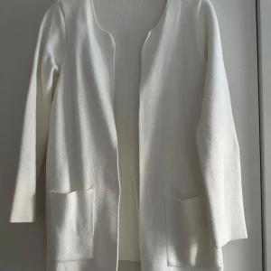 Vit stickad kofta med fickor från Gina Tricot - Säljer en stilren vit stickad kofta med långa ärmar och två stora fickor framtill. Koftan är öppen framtill utan knappar och har en enkel, clean design som passar perfekt till det mesta