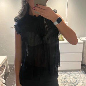 Svart plisserad blus med volang - Säljer en svart blus med plisserat tyg och transparent look. Blusen har volangdetaljer vid axlarna och är ärmlös, vilket ger en cool och trendig vibe. Perfekt för dig som gillar att sticka ut med stilrena detaljer och luftig känsla.