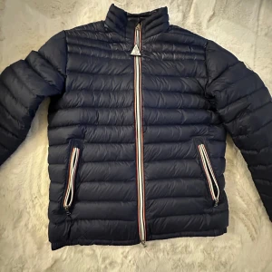 Moncler ”Daniel” dunjacka - Säljer en mörkblå dunjacka från Moncler med klassiska ränder vid dragkedjorna och Moncler-logga på ärmen. Jackan är quiltad, har lång ärm och dragkedja framtill samt vid fickorna. Perfekt för kyliga dagar och riktigt snygg streetstil. Nypris: 13.500kr!! (Och det stiger varje år som går). 