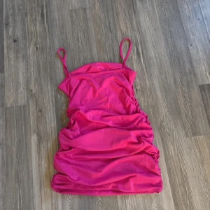 Rosa kort klänning med smala axelband - Säljer en snygg rosa kort klänning med smala axelband och rynkade sidor. Klänningen har en figurnära passform och är perfekt för dig som vill sticka ut. Materialet känns mjukt och följsamt mot kroppen.