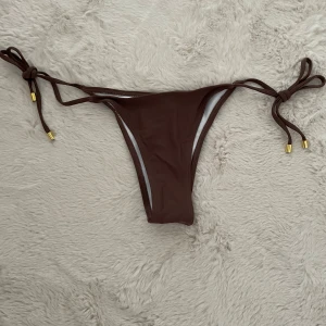 Brun bikiniunderdel  - Snygg brun bikiniunderdel från SHEIN med smal passform och knyt i sidorna med guldfärgade detaljer. Materialet är mjukt och stretchigt syntet, perfekt för strandhäng och poolparty. Enkel och stilren design som ger en fräsch look i sommar.