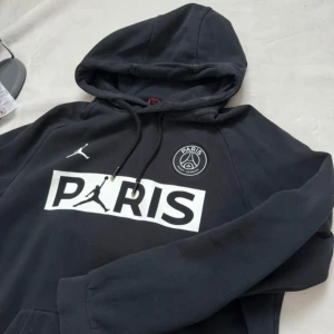 Svart PSG x Jordan hoodie med tryck - Svart hoodie från Jordan x PSG med stor vit PARIS-logga och Jumpman-tryck på bröstet. PSG-emblem på vänster sida och klassisk huva med snörning. Perfekt för dig som gillar streetwear och fotbollsinspirerad stil.