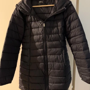 Svart pufferjacka från Only - Säljer en svart  pufferjacka från Only med huva och dragkedja framtill. Jackan har horisontella sömmar, två fickor och är perfekt för kyliga dagar. Den är lätt vadderad och har en stilren look som funkar till det mesta.
