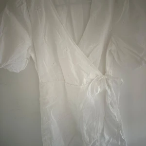 Vit omlottklänning från ONLY - Supersöt vit kortklänning från ONLY med omlottmodell och knyt i midjan. Klänningen har puffiga korta ärmar och små broderade blommor över hela tyget. Perfekt för sommarens alla tillfällen, lätt och luftig i bomull. Tar frakt!