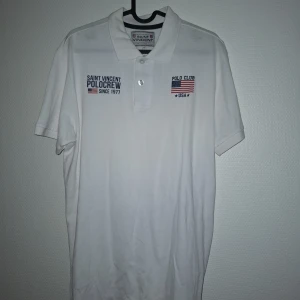 Vit Saint Vincent polo t-shirt M - Vit polo t-shirt från Saint Vincent i storlek M. Klassisk krage med två knappar och broderade detaljer på bröstet med text och flaggor. T-shirten är gjord i mjuk bomull och har korta ärmar. Perfekt för en clean och sportig look.