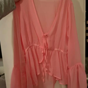 Rosa blus med volang från Pink Woman - Supersöt rosa blus från Pink Woman i luftig polyester med v-ringning och knyt framtill. Blusen har långa ärmar med volang och en flowy, avslappnad passform. Perfekt för dig som gillar romantiska och feminina detaljer.